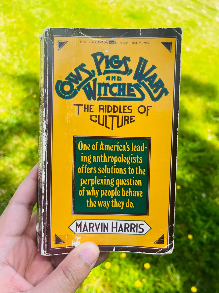 A person holding a physical copy of Cows, Pigs, Wars and Witches by Marvin Harris while reading or reviewing the book.